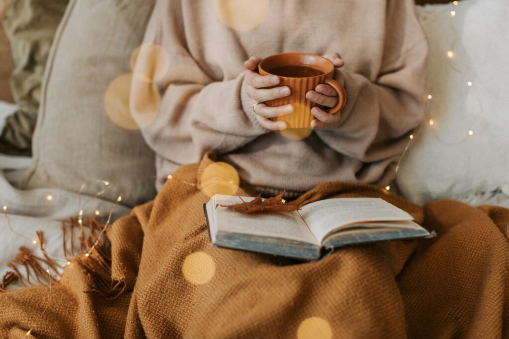 A person sitting indoors holding a warm mug, surrounded by autumn decor, cozy blanket, and book.