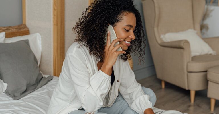 Smiling woman with curly hair working from home on bed with laptop and phone.